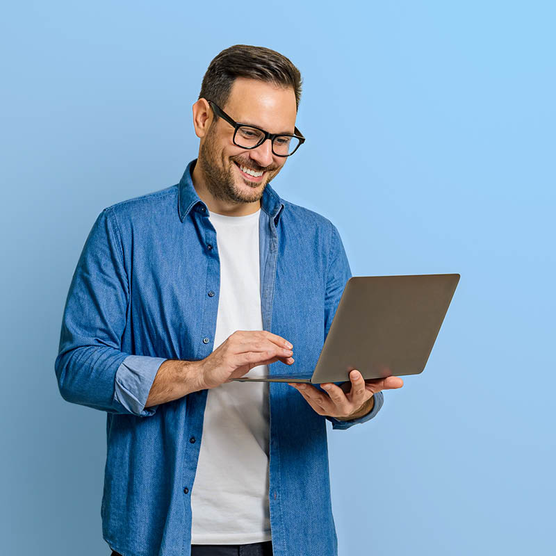 Lächelnder Mann hält einen Laptop und klickt darauf, der Hintergrund ist eine blaue Fläche